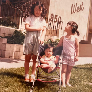 A group of three Korean children stand outside of a Los Angeles apartment building. The youngest child sits in a camping chair.